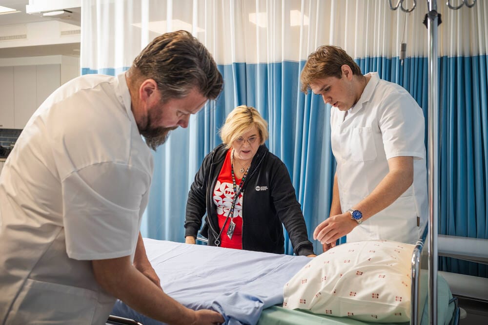 Hoiva-avustajaopiskelijat opettelevat oikeaoppista sängyn sijausta opettajan johdolla.